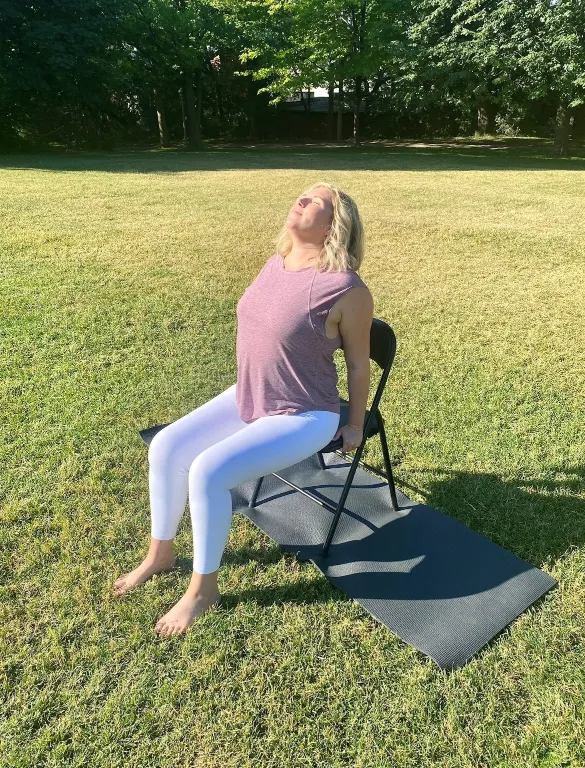 A woman with blonde hair is seated on a folding chair outdoors, performing a gentle chest-opening stretch. She is leaning back slightly, with her hands gripping the back of the chair seat, and her face tilted up toward the sun. She is wearing a maroon tank top and white athletic leggings. The chair is placed on a black yoga mat on a bright, grassy lawn with lush green trees in the background.