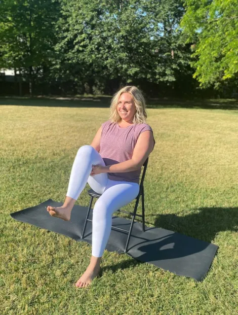 A woman with blonde hair is sitting on a simple folding chair outdoors on a sunny day, demonstrating a gentle exercise or stretch. She is wearing a maroon t-shirt and white athletic leggings. She is seated on a black yoga mat placed on a dry, grassy lawn, with trees and bright sunlight in the background. She is smiling and appears to be lifting one knee towards her chest.