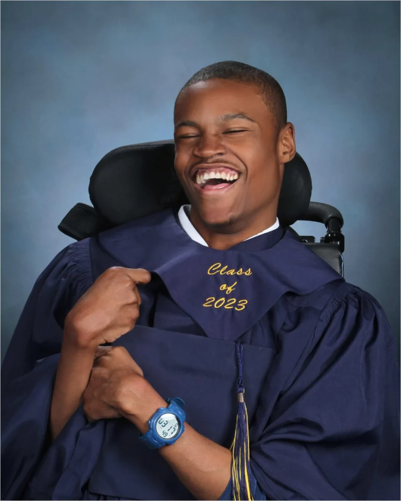 A young Black man in a power wheelchair is wearing a navy blue graduation gown and stole embroidered with "Class of 2023" in yellow cursive text. He is laughing heartily with his eyes squeezed shut and is touching his chest with his left hand. He is wearing a bright blue digital watch. The background is a studio-style blue gradient.