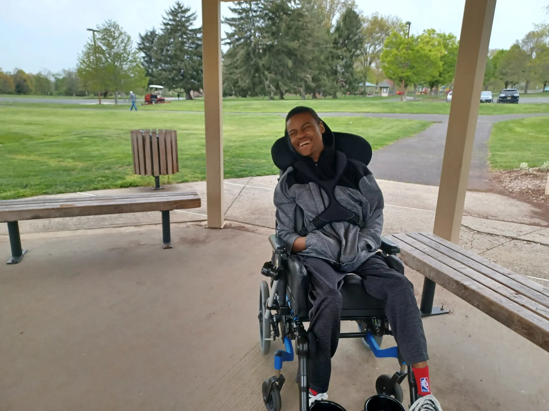 A young Black man is sitting in a power wheelchair under a covered, open-air pavilion. He is smiling widely, wearing a gray zip-up jacket and black pants. He has red socks and white sneakers. In the background, there is a paved pathway, wooden benches, and a large grassy park area with evergreen trees.