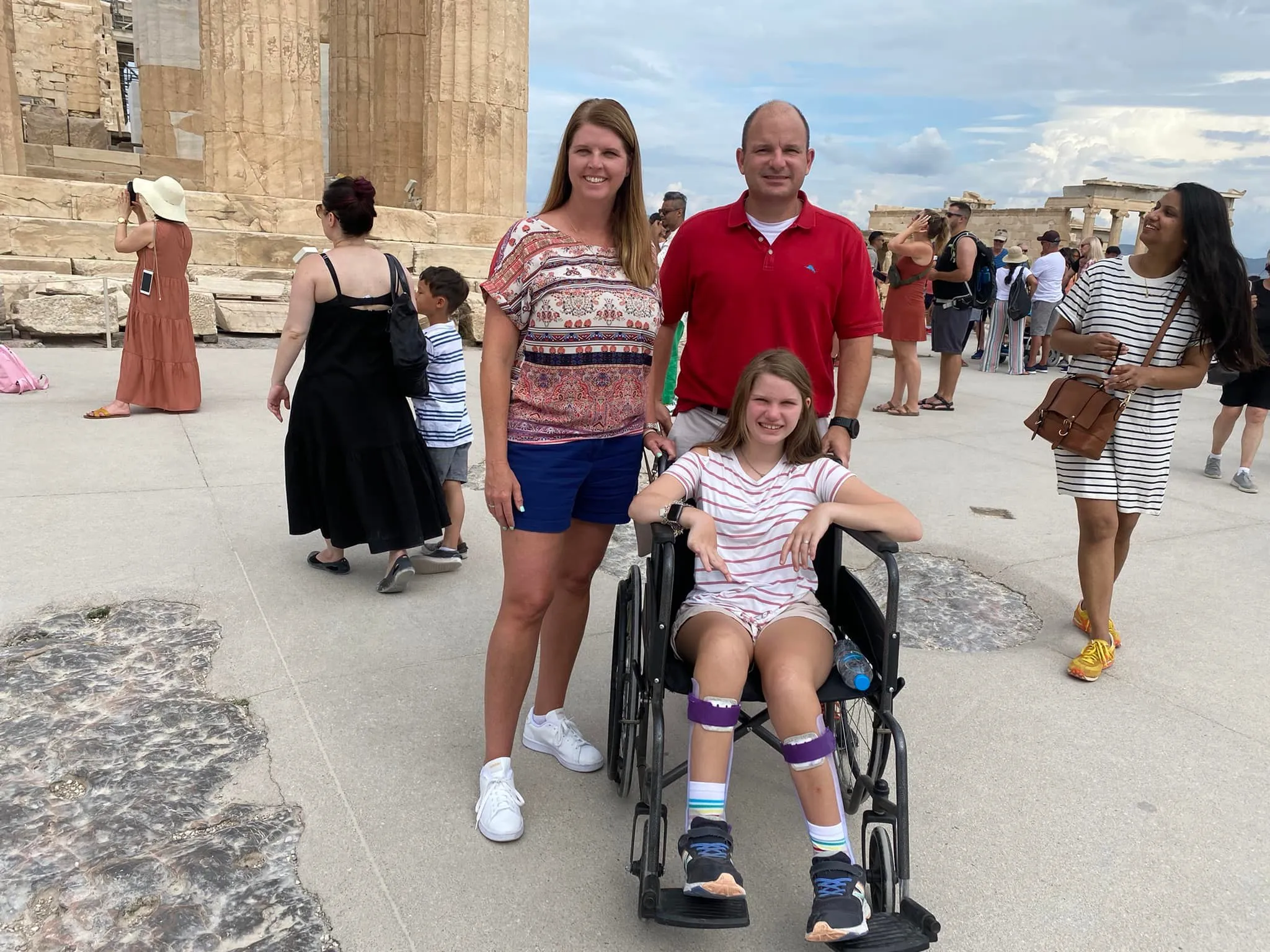 A family of three poses for a photo at a crowded historical site, likely the Acropolis in Athens, Greece, with ancient stone columns visible behind them. The father wears a red polo shirt, and the mother wears a patterned top and shorts. Their daughter is seated in a manual wheelchair, smiling, and wearing a striped t-shirt and shorts with braces or supports on her lower legs and ankles. Tourists are visible all around them in the background.