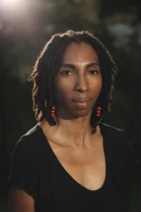 The image shows a person, Saada Branker, with medium-length, dark dreadlocks, wearing a black top. They have a calm, serious expression, and their face is highlighted by soft lighting. The background is slightly blurred, making the person the focal point of the image. They are also wearing earrings with red and yellow accents, which stand out against the dark backdrop. The overall mood of the image feels contemplative and serene.
