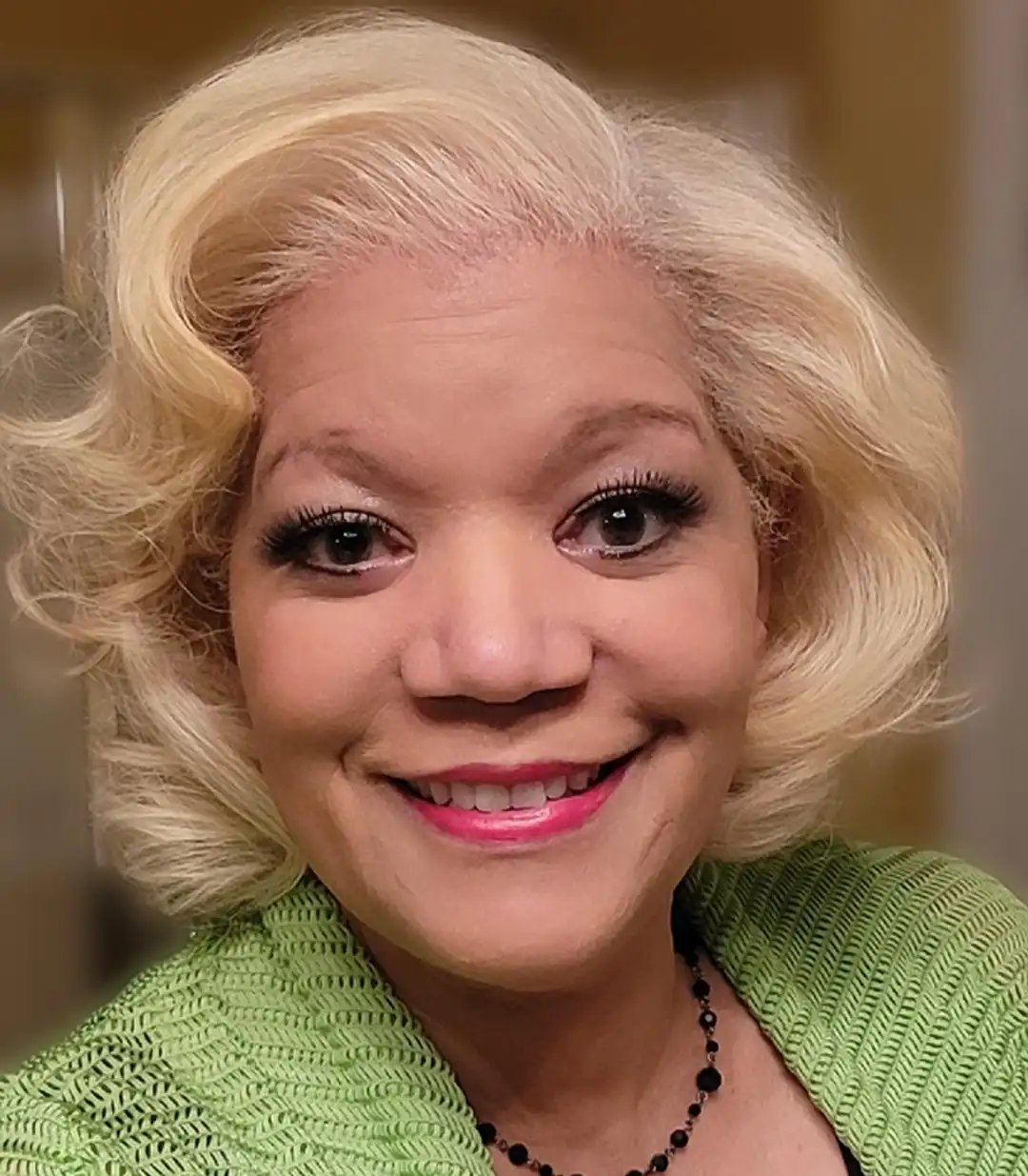 A close-up headshot of a smiling woman with short, wavy, platinum blonde hair. She is wearing a bright pink lipstick, dark eyeliner and lashes, and a lime green textured sweater or cardigan. She is also wearing a black beaded necklace. The background is softly blurred and neutral-toned.