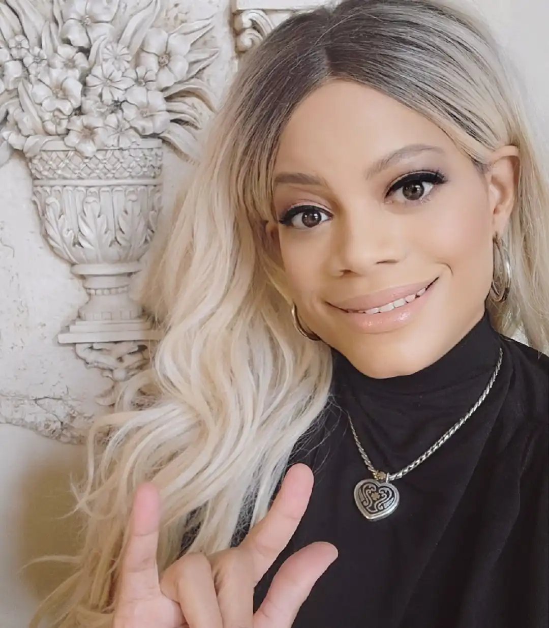 A smiling woman with long, wavy blonde hair and a black turtleneck top is taking a selfie indoors. She is wearing a silver heart locket necklace and silver hoop earrings, and she is holding her hand up in the "I love you" sign. A decorative white floral relief on a wall serves as the background.
