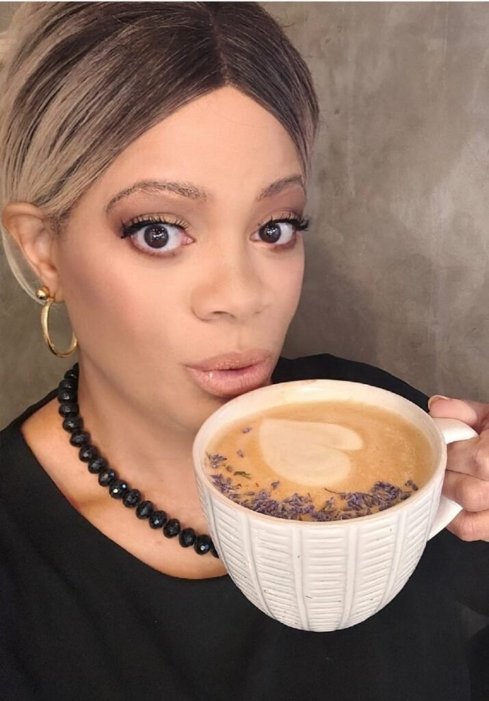 A woman with blonde-highlighted hair, wearing a black top and a black beaded necklace, holds a white mug filled with a latte. The latte has a heart shape made of foam on top and is garnished with small purple sprinkles or herbs (possibly lavender). She is looking directly at the camera with a surprised or curious expression.