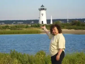 A woman with curly brown hair, wearing a light-colored t-shirt and dark pants, stands outdoors next to a body of water. She is looking at the camera and pointing toward a distant, white, conical lighthouse with a black top that stands beyond the water and a grassy bank. A few houses and a sailboat mast are visible in the far background. The lighthouse appears to be the Edgartown Lighthouse on Martha's Vineyard.