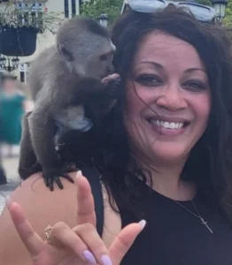 A smiling woman with long dark, curly hair wearing a black top and sunglasses resting on her head. A small capuchin monkey is sitting on her left shoulder, appearing to feed itself. The woman is holding up her left hand in a "rock on" sign (thumb, index, and pinky fingers extended) and is wearing a ring. The background is slightly blurred, showing some outdoor structures.
