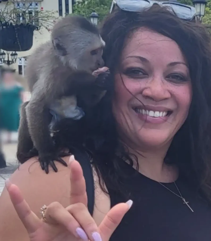 A close-up selfie of a smiling woman with long, dark, wavy hair. A small, brown and beige capuchin monkey is sitting on her right shoulder, appearing to eat something it holds in its hands. The woman is wearing a black top and a cross necklace, and she holds her left hand up in the "I love you" sign. The background is blurred, suggesting an outdoor, possibly public or historical setting.