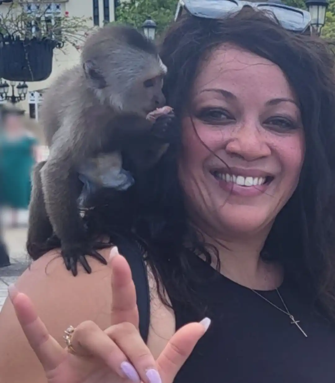A close-up selfie of a smiling woman with long, dark, wavy hair. A small, brown and beige capuchin monkey is sitting on her right shoulder, appearing to eat something it holds in its hands. The woman is wearing a black top and a cross necklace, and she holds her left hand up in the "I love you" sign. The background is blurred, suggesting an outdoor, possibly public or historical setting.