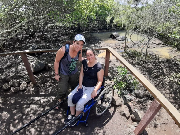 Two women outdoors on a dirt path next to a wooden railing. One woman is standing, and the other is seated in a blue off-road wheelchair. The background shows dense green foliage and a muddy area where three large tortoises are visible, one near the women and two partially submerged in a puddle.