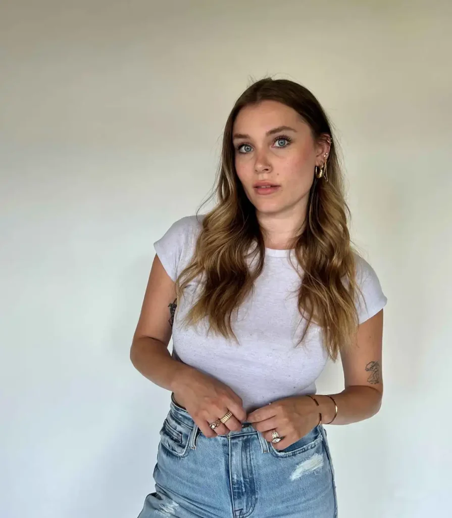 A young woman with long, wavy brown hair and blue eyes stands against a plain white background. She is wearing a fitted white cap-sleeve t-shirt and light-wash denim jeans with a distressed or ripped detail. She wears gold hoop earrings, a wrist bracelet, and several gold rings, and she is looking directly at the camera.