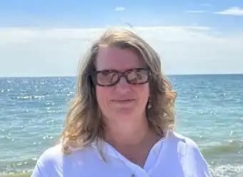 A middle-aged woman with light brown, wavy hair and tortoiseshell glasses is smiling faintly while wearing a white shirt. She is standing outdoors on a bright day, directly in front of a sparkling blue ocean or large lake, with the water filling the background.