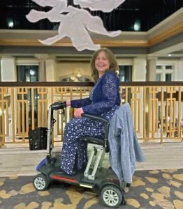 A smiling middle-aged woman with long brown hair is seated on a black and red motorized mobility scooter indoors. She is wearing a dark blue outfit with a white floral or patterned design, including a lace overlay on the top. A gray jacket is draped over the back of the scooter. The setting appears to be a large, elegant hotel lobby with high ceilings, decorative white lighting suspended above, and an ornate carpet.