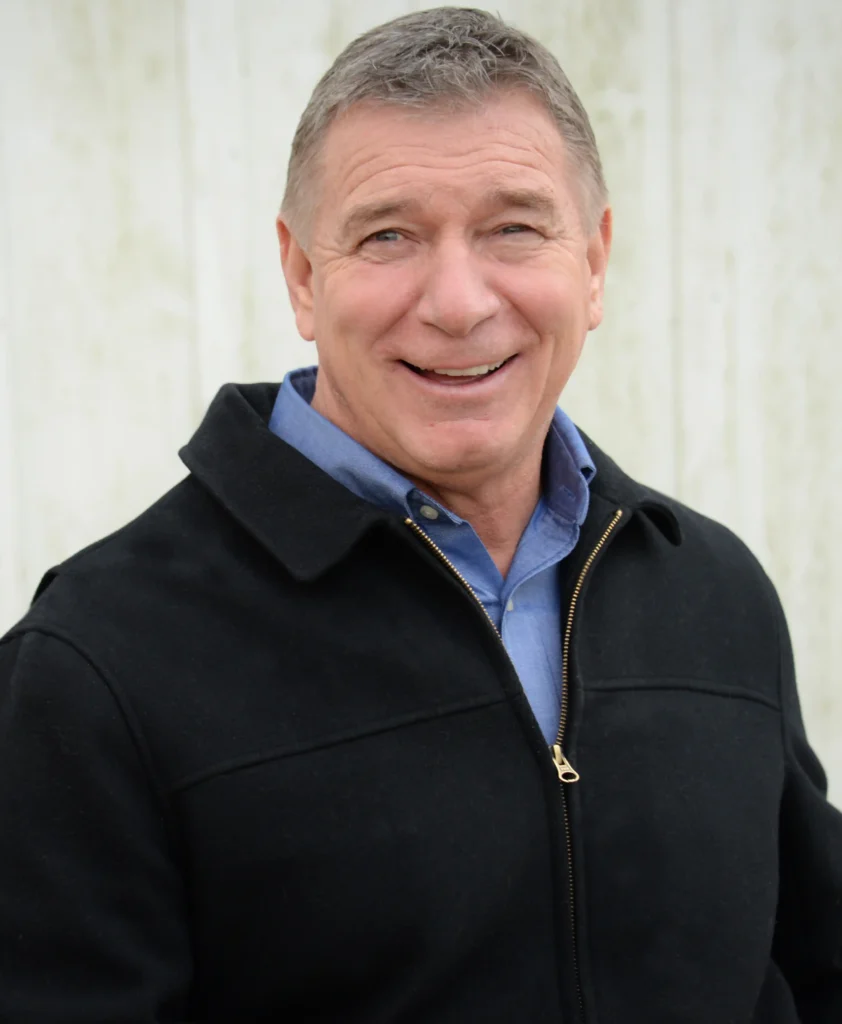 A head-and-shoulders portrait of a middle-aged man with short, graying hair, smiling at the camera. He is wearing a bright blue collared shirt beneath a black zip-up jacket. The background is a plain, lightly colored, vertically paneled surface.