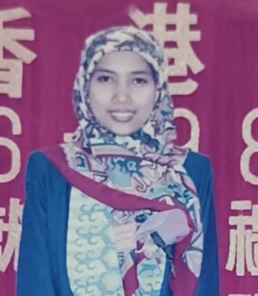 A young woman is smiling and wearing a hijab made of a multicolored, patterned scarf with paisley and geometric designs, along with a blue top. She is standing in front of a bright red banner or curtain that has large, yellow or white Chinese characters on it.