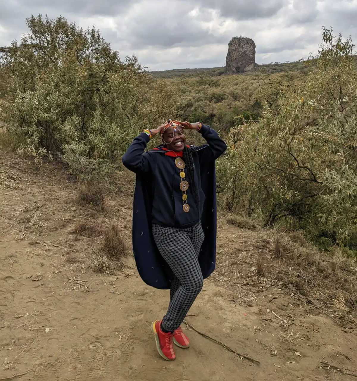 A person with facial paint, wearing a dark blue hoodie, a long navy cape, checkered grey pants, and bright red shoes, stands on a dry dirt path outdoors. They are smiling and holding their hands up to their head. In the background, there is dense, dry shrubbery and a distinctive, large, rocky spire or formation rising up from the landscape under a cloudy sky. The setting appears to be a dry, bushland environment, possibly in Kenya near a feature like the Pride Rock inspiration.