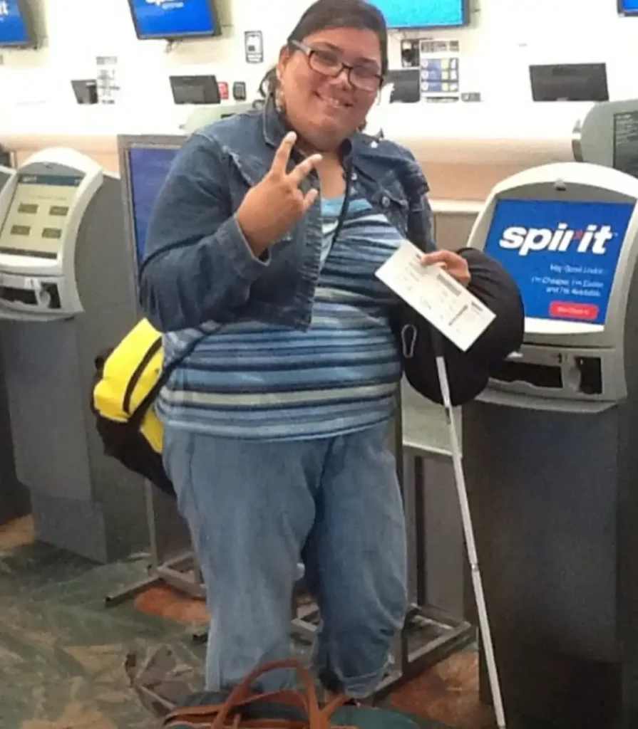 A smiling woman with dark hair and glasses stands in an airport check-in area. She is wearing a denim jacket, a blue striped shirt, and blue jeans, and she is holding a white cane and a piece of paper that appears to be a boarding pass or ticket. She is holding up her left hand in a peace sign. Two Spirit Airlines self check-in kiosks are visible on either side of her, and she has a backpack with yellow straps resting on the floor.