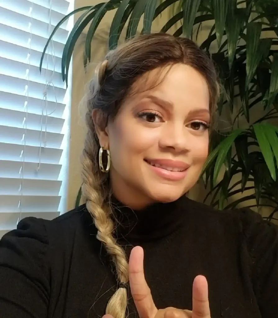 A woman with blonde-highlighted hair styled in a side braid, wearing a black turtleneck top, is smiling indoors. She is holding her right hand up in the "I love you" sign. The background includes a large green houseplant and white window blinds.