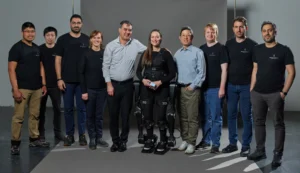 The image shows a group of ten individuals posing together for a photograph. They are standing in a line with a grey backdrop, suggesting a formal team portrait setting. The group is diverse in gender and ethnicity. One notable feature is that the fifth person from the left is wearing an exoskeleton mobility device. The shirts worn by seven individuals have the same logo, representing the Human in Motion Robotics team. 