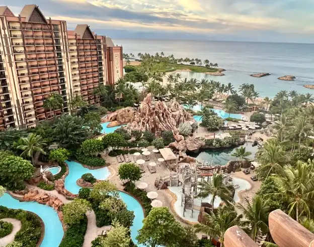 An aerial view of a luxury tropical resort during early evening. The resort is composed of a large, multi-story building with a terracotta roof and balconies, suggesting a design inspired by the local architecture. In the foreground, a meticulously designed pool area mimics a lazy river, winding through an artificial landscape of rocks and palm trees. The pool area is dotted with lounge chairs and umbrellas for relaxation. Beyond the resort, the calm ocean extends to the horizon under a soft, pastel sky, indicating the resort's beachfront location. The tranquil setting is designed for leisure and suggests an atmosphere of an exclusive getaway.