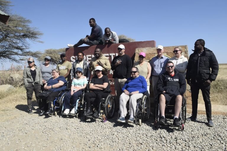 A diverse group of people posing for a photo at an outdoor location, which appears to be a desert or savannah setting. Several individuals are seated in wheelchairs, suggesting the group may have a focus on accessibility or inclusivity in travel or outdoor activities. There's a sign behind the group, partially obscured, which might indicate the name of the location. The mood is casual and it seems like a bright, sunny day. There's an array of different clothing styles, indicating varying personal tastes and perhaps a range of temperatures. Overall, it's a moment of camaraderie and shared experience.