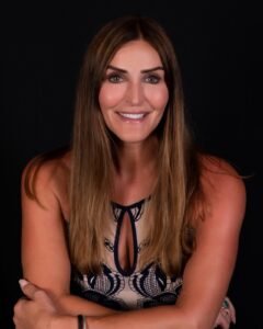 A woman with long, light brown hair and a healthy tan is smiling at the camera. She has striking blue eyes and is wearing a sleeveless navy blue and white patterned dress with a plunging neckline featuring a black detail. Her arms are crossed in front of her, and one hand is adorned with a large green ring. The background is plain black, emphasizing her and the bright colors of her outfit.