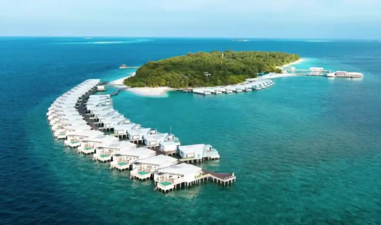 A panoramic view of a luxurious water villa resort located in the Maldives. The villas are arranged in a curving line, extending into the azure sea. Each unit has direct access to the water, with some featuring private pools. A lush, green island forms the backdrop, with additional resort facilities and possibly a beach. The clear blue skies and calm seas suggest an idyllic, serene vacation spot.