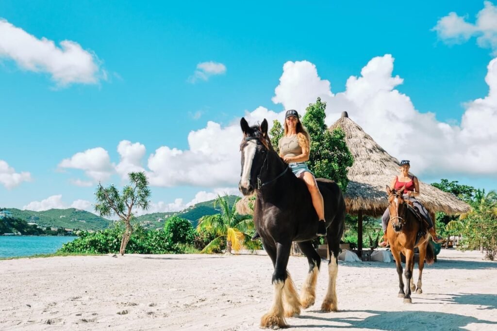 Two women enjoying a sunny day horseback riding on a tropical beach. The woman in the foreground is on a large black horse with prominent fluffy fetlocks, smiling towards the camera. She's wearing a cap, a light-colored tank top, and shorts. The second woman, in the background, is riding a smaller brown horse and is also smiling, dressed in a pink top and dark pants. They are riding on sandy ground, with lush green hills, palm trees, and a traditional thatched-roof structure in the background, under a bright blue sky dotted with fluffy clouds.