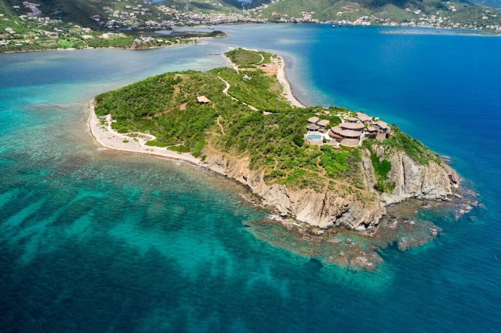 An aerial view of a lush, green peninsula jutting into clear blue waters. Several upscale buildings with thatched roofs are scattered along the ridge, enjoying expansive views of the surrounding sea. The coastline features a mix of rugged cliffs and sandy beaches, providing a natural boundary for this secluded retreat. To the left, a calm bay with boats anchored near the shore is visible, while the right side showcases the open ocean, hinting at a vibrant marine ecosystem below the surface.