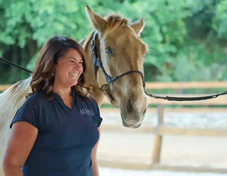 A woman smiling broadly and standing to the left with her body angled slightly towards the camera. She has long brown hair, is wearing a dark blue t-shirt with a logo on the left side, and appears to be enjoying a moment with a horse. The horse, positioned on her right, has a sandy blonde coat and is wearing a blue halter. The background suggests an equestrian setting, possibly an arena, with trees and a fence visible in the distance.