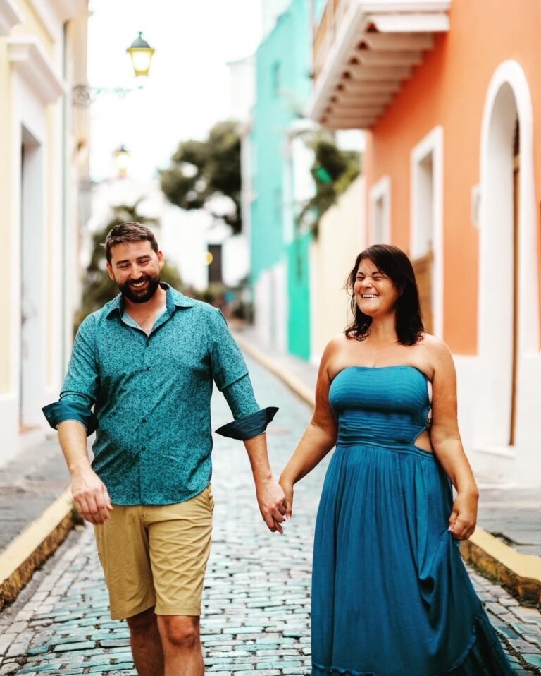 A couple holding hands and smiling as they walk down a cobblestone street. The man is on the left, wearing a teal printed shirt and beige shorts. The woman is on the right, wearing a strapless teal dress. The street is lined with colorful buildings and a lantern is visible in the background. They appear happy and relaxed, enjoying their stroll.