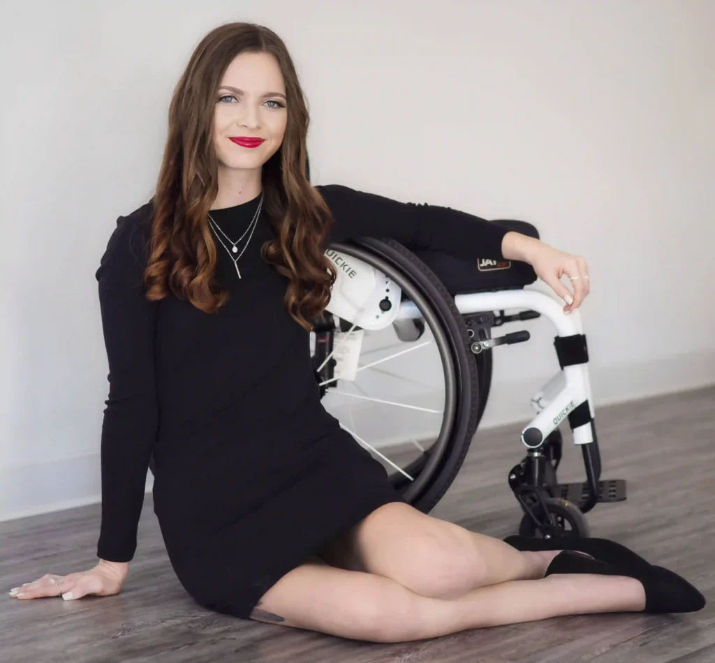 A woman sitting on the floor next to a wheelchair, smiling at the camera. She has long, wavy brown hair and is wearing a black dress, black flats, and red lipstick. She has a necklace on, and her left hand is resting on the floor while her right hand is on the wheel of the wheelchair. The background is a plain, light-colored wall, giving a clean and simple aesthetic to the photo.