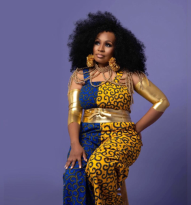 A woman posing confidently, wearing a vibrant blue and yellow patterned outfit with golden accents. She has voluminous black curly hair, large hoop earrings, and multiple necklaces. Her attire includes a gold arm cuff on her left arm and she's adorned with a matching gold waist belt. The background is a plain purple, which complements her attire.