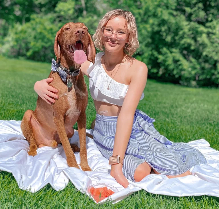 A young woman and her brown dog are enjoying a sunny day outdoors, sitting on a white blanket spread over lush green grass. The woman, sporting blonde, shoulder-length hair with glasses, is dressed in a summery, white cropped top and blue striped skirt. She's smiling gently at the camera. The dog is looking content with its tongue out has a golden-rust color. They both seem to be relishing a perfect day in the park.