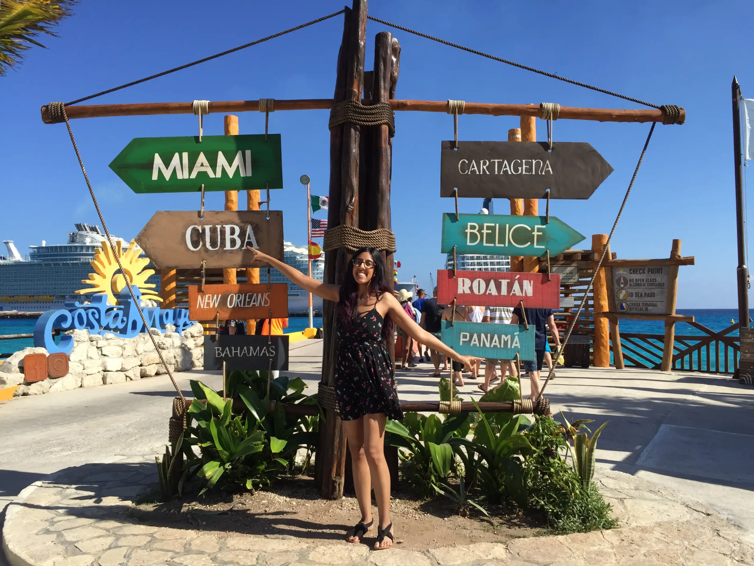 A woman stands smiling at a crossroads signpost in a sunny, tropical location. The signpost includes directions to various destinations like Miami, Cuba, New Orleans, Bahamas, Cartagena, Belize, Roatan, and Panama. In the background, the clear blue sky complements the bright blue sea where a cruise ship is docked, hinting at a popular tourist port, possibly Costa Maya in Mexico, as suggested by another sign. The woman is dressed in a summer outfit suitable for vacationing, with sunglasses and a hat that suggests a relaxed holiday vibe.