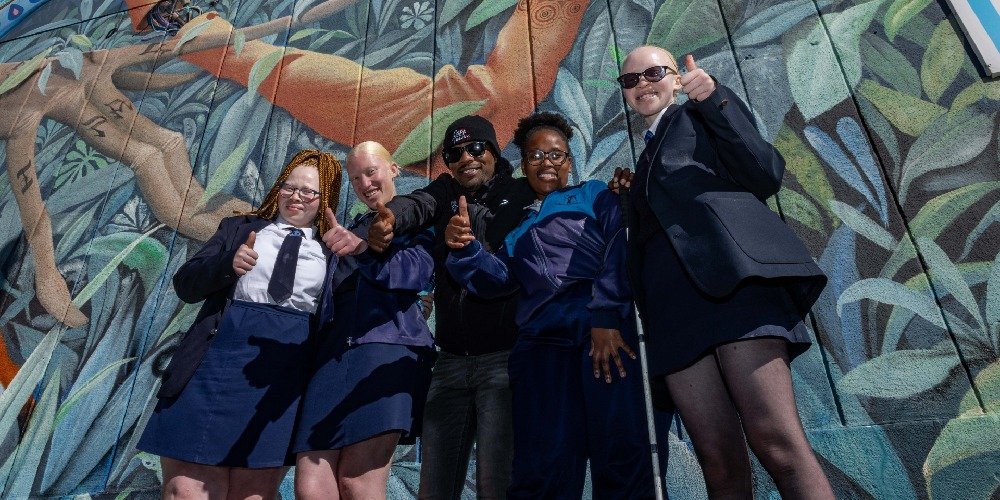 A group of four people in high spirits posing in front of a vibrant mural. They are dressed in professional attire, with two wearing jackets and ties, and the others in sporty windbreakers. The mural behind them depicts an abstract, nature-inspired scene with green leafy patterns. The group seems to be in a celebratory mood, showing thumbs-up and peace signs to the camera, reflecting a positive and cohesive atmosphere.