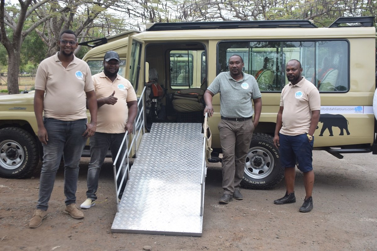 Four men stand in front of a safari vehicle in a natural, outdoor setting. They are wearing matching polo shirts with a logo on the left chest, possibly indicating they are part of a team or organization. The safari vehicle is equipped with a foldable ramp, suggesting that it may be used for accessible tourism or transport of equipment. The vehicle is painted a light tan color, a common choice for safari tours to blend with the environment and for better viewing of wildlife. The men seem to be in a relaxed pose, likely signifying a moment of rest or preparation before or after an activity.