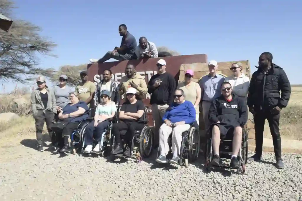 A diverse group of people posing for a photo at an outdoor location, which appears to be a desert or savannah setting. Several individuals are seated in wheelchairs, suggesting the group may have a focus on accessibility or inclusivity in travel or outdoor activities. There's a sign behind the group, partially obscured, which might indicate the name of the location. The mood is casual and it seems like a bright, sunny day. There's an array of different clothing styles, indicating varying personal tastes and perhaps a range of temperatures. Overall, it's a moment of camaraderie and shared experience.