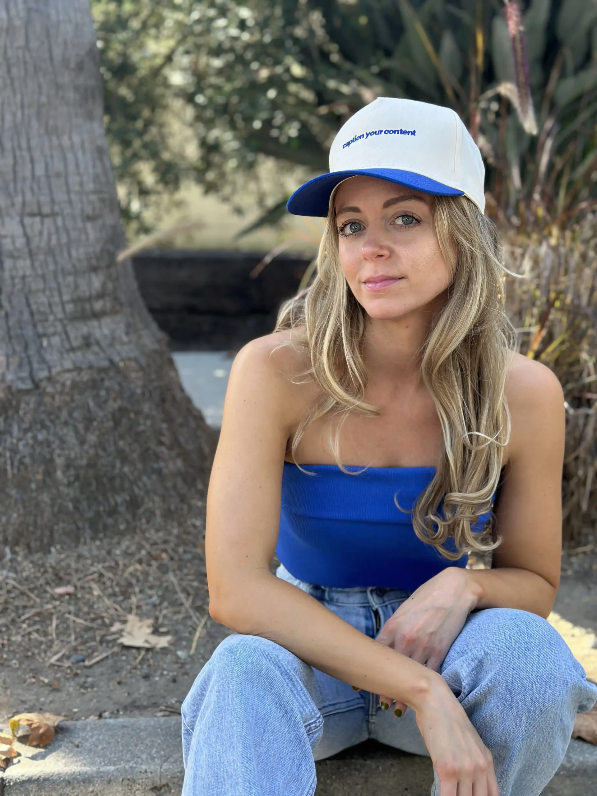 A woman sitting outdoors with a casual pose. She's wearing a blue tank top, light-wash denim jeans, and a white and blue baseball cap with the text "caption your content" on it. Her blonde hair is styled in loose waves, and she looks contemplative or relaxed. In the background, there's a tree and some shrubbery, indicating a park or a similar green space.