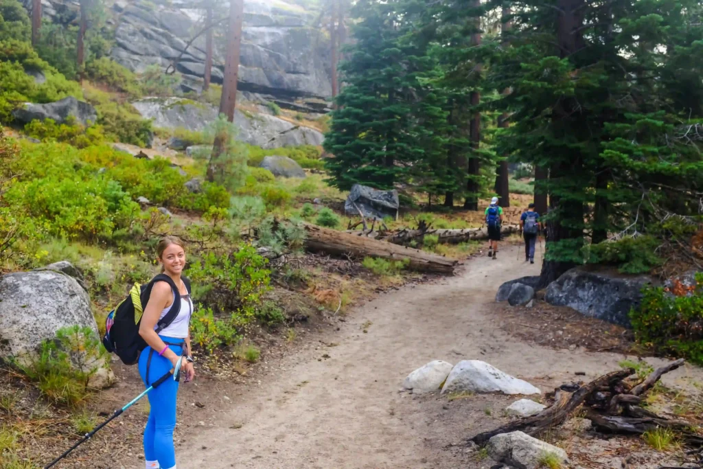 A woman with a cheerful expression looks back towards the camera on a forest hiking trail. She is equipped with a walking pole and a backpack, suggesting she's prepared for an extended trek. The trail appears well-trodden, bordered by rocks, green shrubbery, and towering pine trees. In the background, two other hikers with backpacks are walking away from the camera, further along the path, which suggests this is a popular route for nature enthusiasts. The overcast sky indicates potential cool or early morning weather, ideal for hiking.