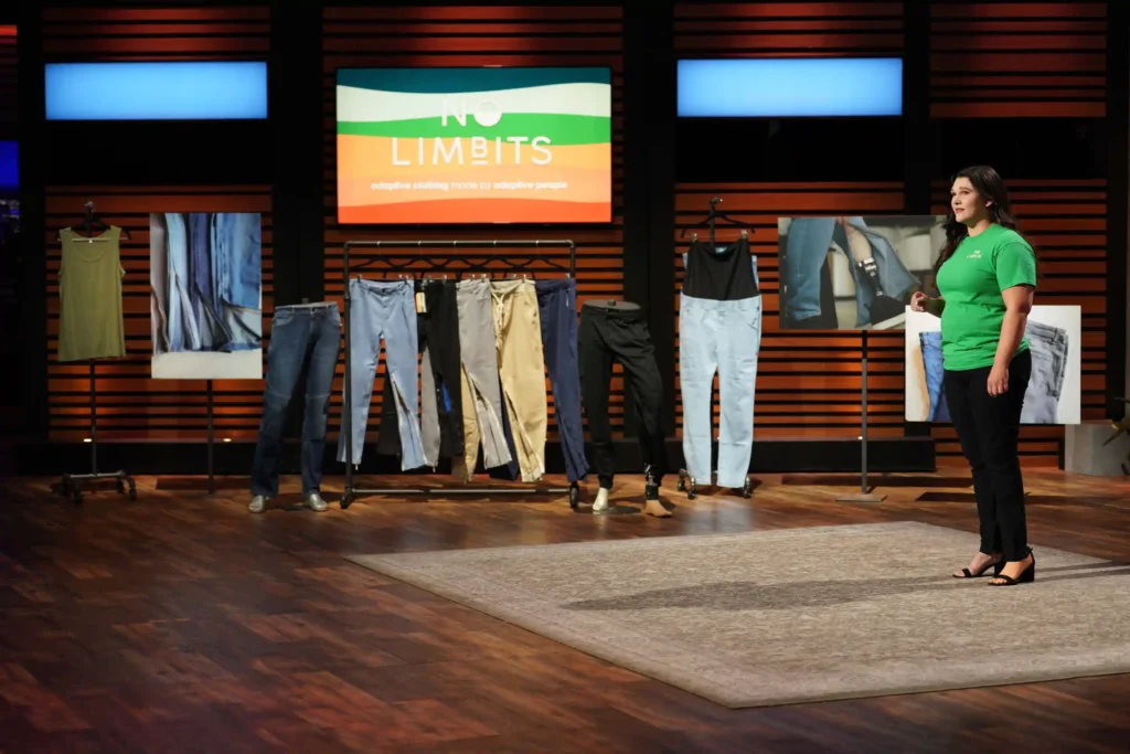 A woman standing on a stage next to a rack of different styles of pants displayed on stands. Behind her is a large screen with the text "NO LIMBITS adaptive clothing." On the left, there is a single piece of clothing on a stand, a pair of pants. The stage has wood paneling and spotlights with an overall professional appearance, indicative of a business or product pitch environment, reminiscent of television shows where entrepreneurs present their products to potential investors.