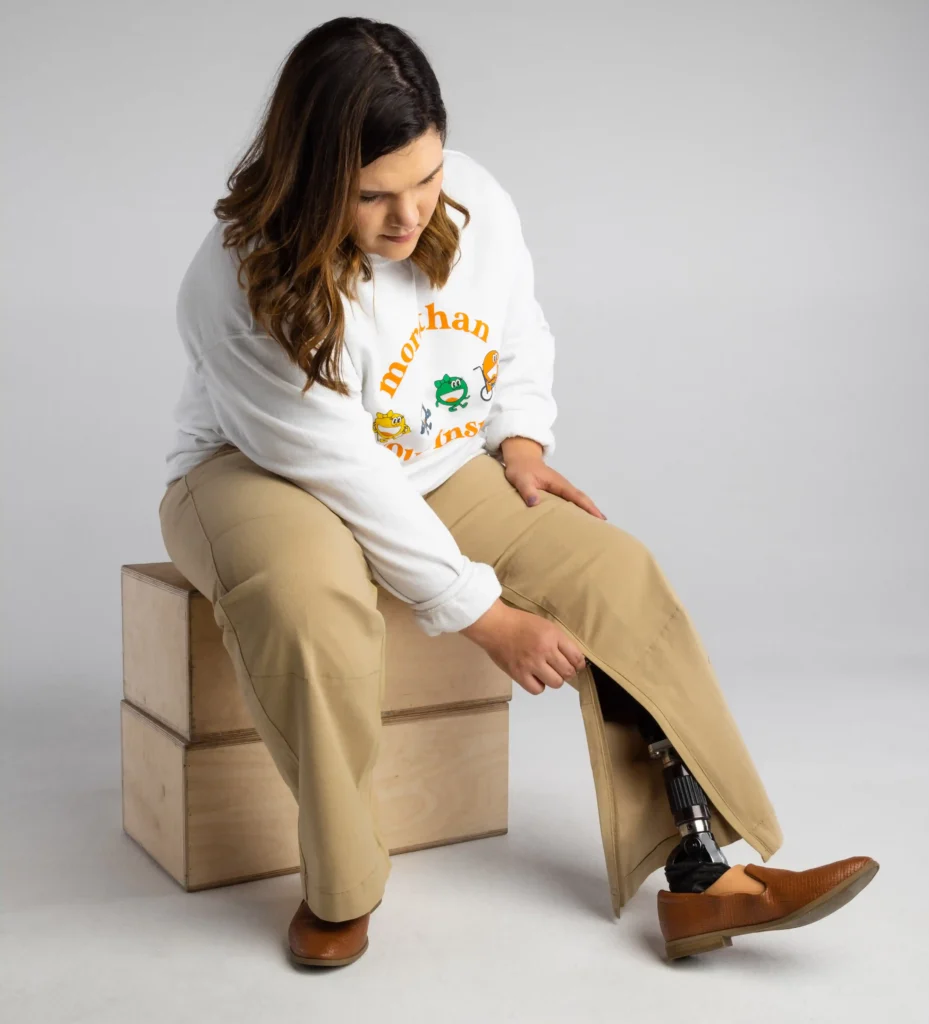 A person with a prosthetic lower leg is seated on a wooden box, looking down at their prosthetic limb as the pull the zipper down on heir pants to cover the prosthetic leg. They are dressed casually in khaki pants and a white sweatshirt with a graphic design featuring cartoon frogs and the text "More than Limbs". The individual is wearing one brown shoe, and the background is plain and light-colored, providing a neutral setting that highlights the subject.