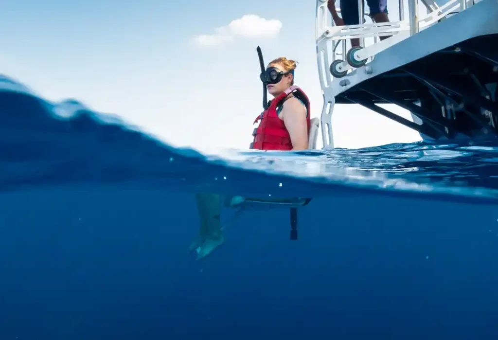 A disabled person in a red life vest and black snorkeling mask sits on the edge of a boat, preparing to enter the ocean. Half of the image shows the scene above water, where we see the boat and the person, and the other half is underwater, offering a view of the boat's submerged part and the clear blue sea.