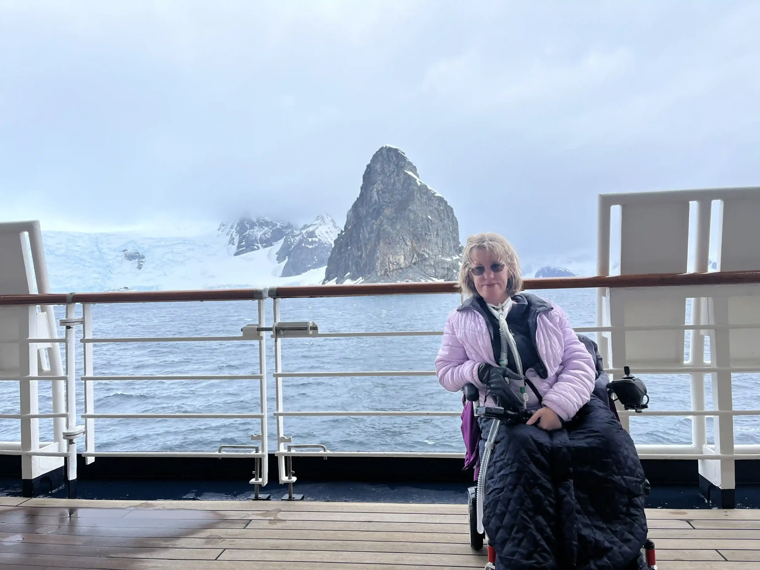 A woman in a wheelchair on the deck of a ship, bundled up in a lilac puffer jacket and wearing sunglasses. Behind her, the vast, tranquil sea stretches out to the horizon, meeting a sky partially veiled with soft clouds. To her right, the deck railing provides a clear view of a rugged, snow-covered mountain peak in the distance, evoking a sense of adventure and the serene beauty of a cold, maritime landscape.
