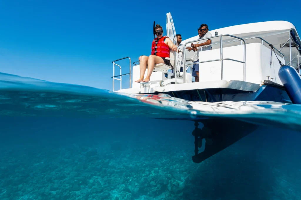 A split-view image showing the sunny, clear sky above and the crystal-clear blue ocean below. On the boat deck, a person with a disability wearing snorkeling gear and a red life vest sits on the edge, ready to enter the water. Another person stands behind the helm, steering the boat. A third individual appears to be preparing for the water activity. The underwater part of the boat is visible, with the sea floor beneath, hinting at the clarity and shallowness of the water.