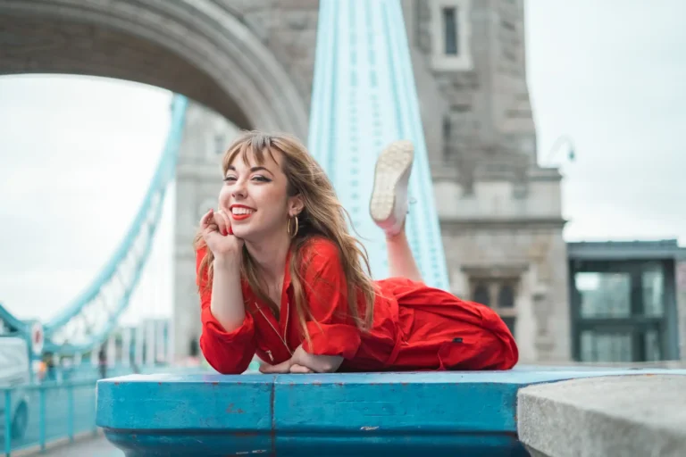 A woman in a bright red jumpsuit is lying on her side on a blue bridge railing with the Tower Bridge in the background. She is smiling with her head propped up by her hand, and one leg playfully kicked up in the air. Her joyful expression and the iconic London landmark suggest a carefree, tourist moment captured on camera.