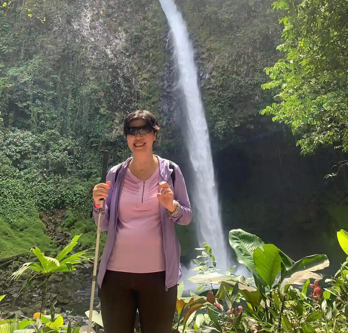 A blind lady stands smiling in the foreground, wearing sunglasses, a light purple top with a darker purple zip-up jacket, and dark pants. They hold a walking stick in one hand and give a thumbs-up with the other. Behind them, a majestic waterfall cascades down a rocky cliff amidst lush green vegetation. Sunlight filters through the tree leaves above, adding a bright, natural glow to the serene landscape.