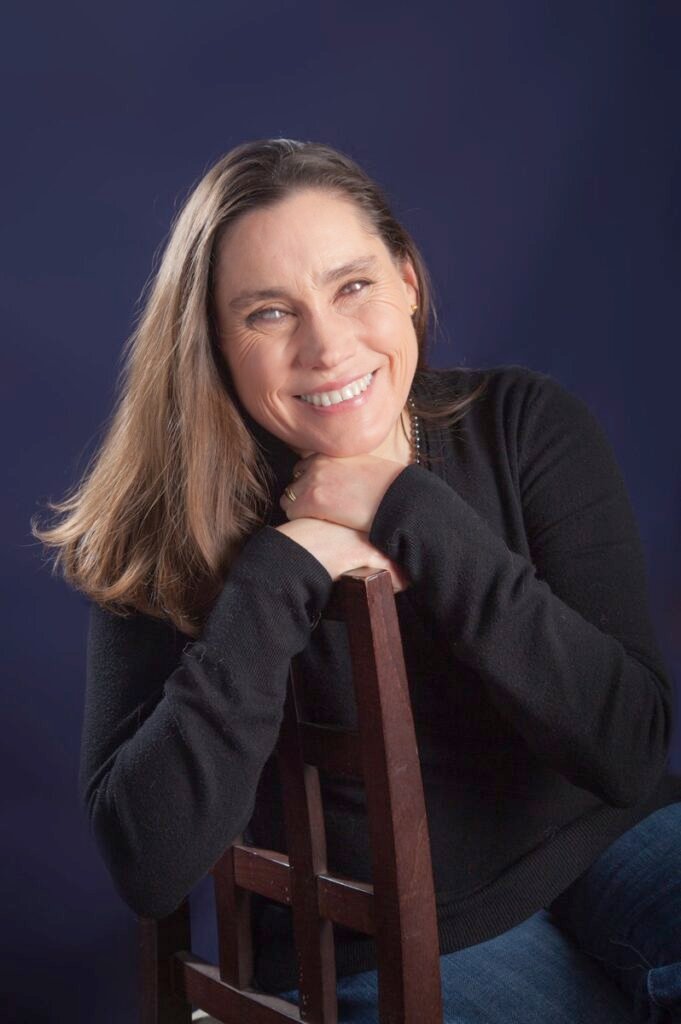 A blind woman, Lois-Strachan, leaning on a wooden chair, smiling towards the camera. She has long brown hair, is wearing a black sweater and denim jeans, and has a warm, friendly expression. The background is a solid dark blue, providing a nice contrast with the subject, which gives the portrait a classic and clean appearance.