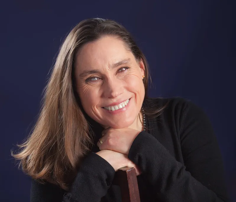 A blind woman, Lois-Strachan, leaning on a wooden chair, smiling towards the camera. She has long brown hair, is wearing a black sweater and denim jeans, and has a warm, friendly expression. The background is a solid dark blue, providing a nice contrast with the subject, which gives the portrait a classic and clean appearance.