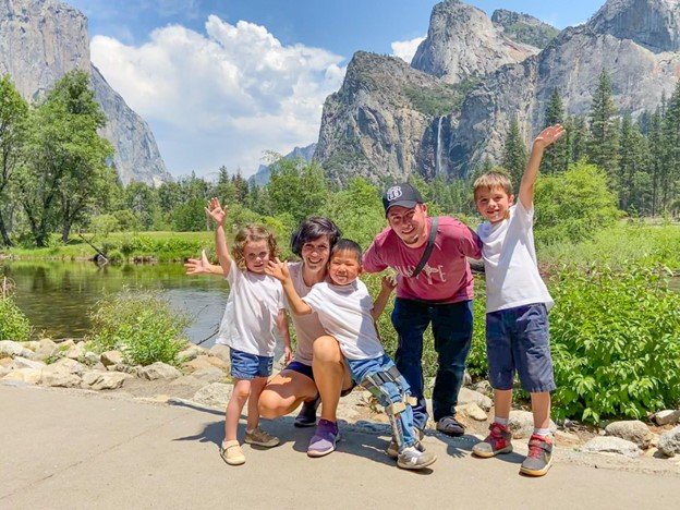 A group of five people posing happily for a photograph with a scenic backdrop of majestic mountains and a serene river. Two adults are crouched down with three children, each person is smiling broadly and some of the children have their arms raised in excitement. They're enjoying a sunny day outdoors in a picturesque valley, likely a national park, surrounded by the natural beauty of towering cliffs and lush greenery reflected on the calm water surface.